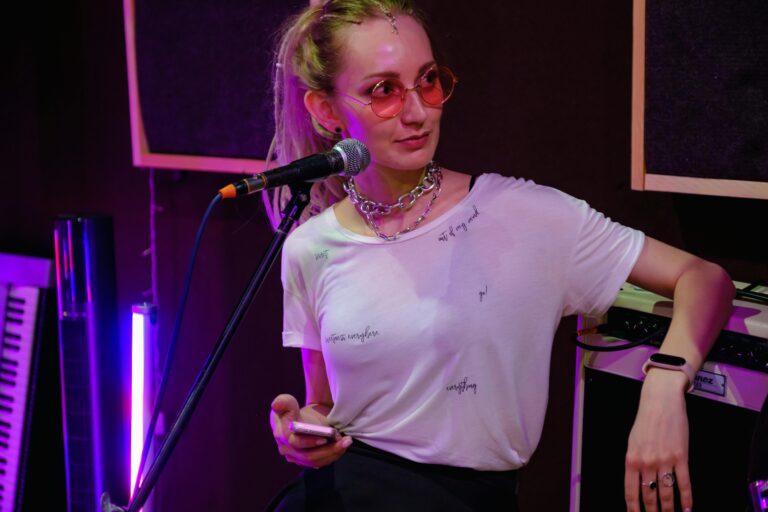 Femme avec lunettes roses, micro et clavier lumineux en arrière-plan.