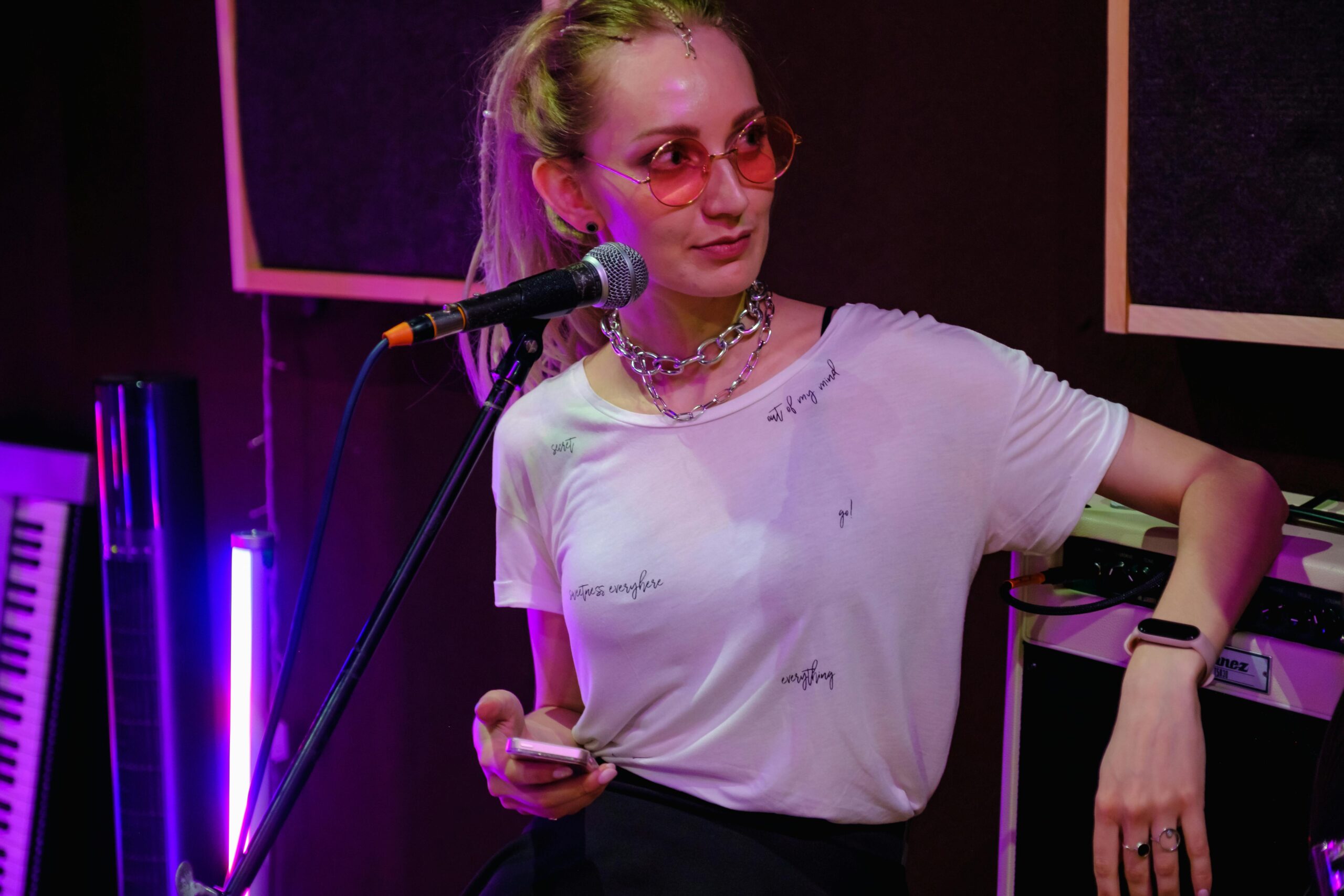 Femme avec lunettes roses, micro et clavier lumineux en arrière-plan.